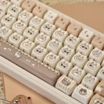 This image shows the charming Azukisan Siamese Cat PBT keycaps set from a playful angle, highlighting the whimsical cream and brown stylized cat motif.