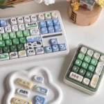 Cow pasture keycaps MOA PBT dye-sub set shown on a keyboard and matching number pad, with cream cows, green meadows, blue sky, and milk accents.