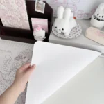 Hand lifting the smooth white back of a pastel pink lolita cat-girl kawaii anime desk mat, shown waterproof and non-slip beside cute plush desk decor.