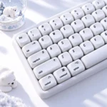 Close-up of white Polar Bear keycaps from a snowy MOA 139-key PC set, with a snow globe and sleeping bear figurine adding cozy winter charm.